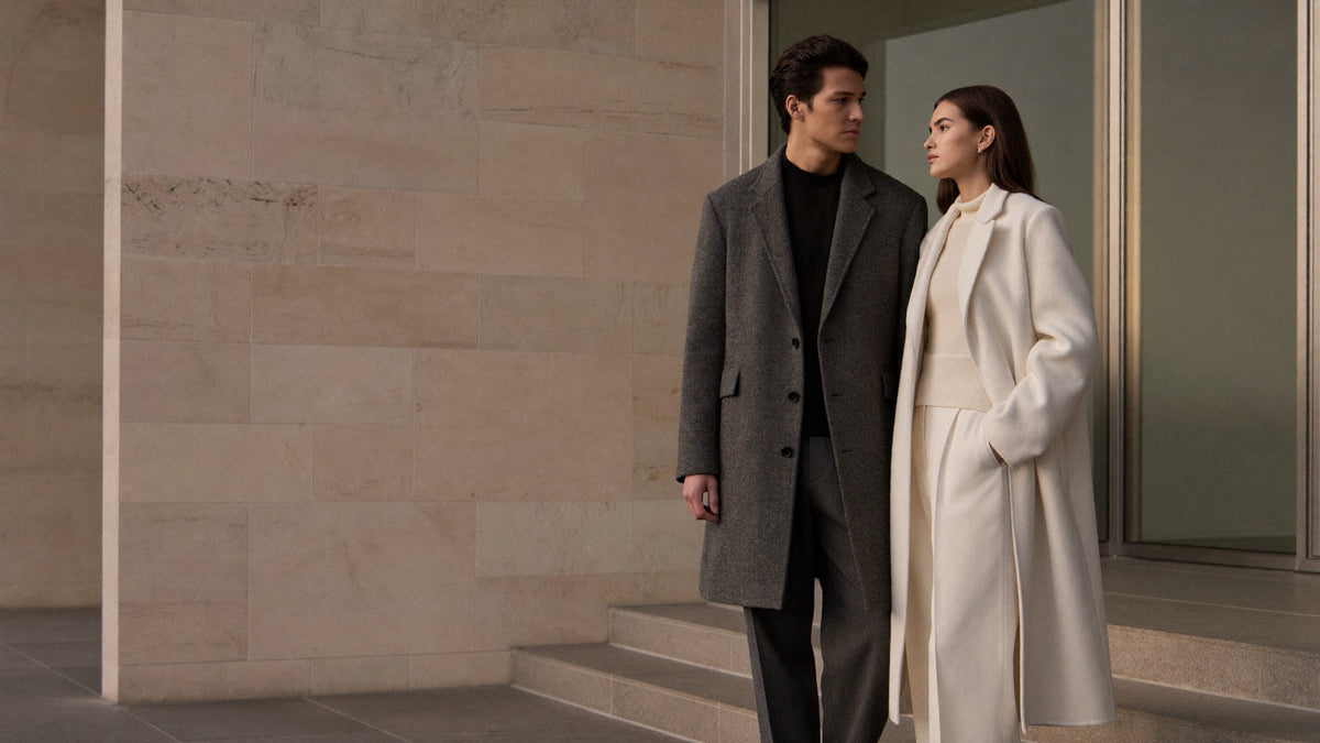 A man in a charcoal overcoat and a woman in an ivory coat standing by a stone building